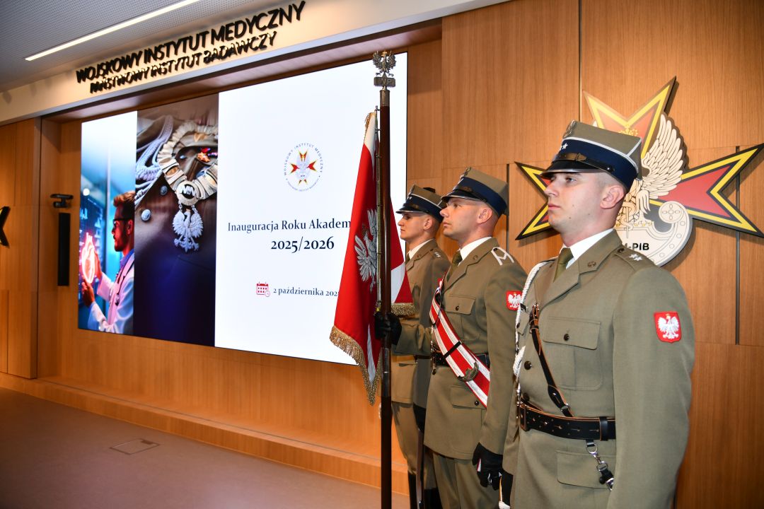 Aula WIM-PIB, trzech żołnierzy w mundurach galowych stoi na baczność z sztandarem Polski, obok nich wyświetlony napis Inauguracja Roku Akademickiego 2025/2026.