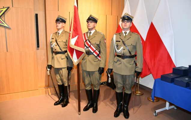 Trzech żołnierzy w mundurach galowych stojący na baczność. Żołnierz po środku trzyma sztandar.