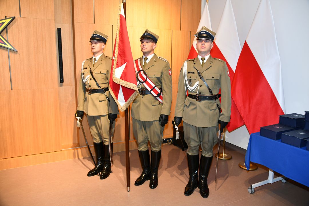 Trzech żołnierzy w mundurach galowych stojący na baczność. Żołnierz po środku trzyma sztandar.