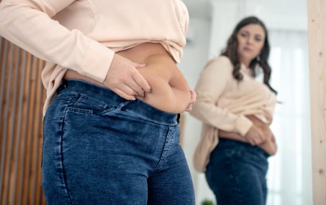 Kobieta z nadwagą przeglądająca się w lustrze, trzyma fałdę brzucha.