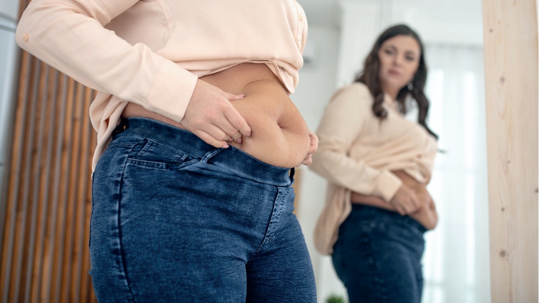 Kobieta z nadwagą przeglądająca się w lustrze, trzyma fałdę brzucha.