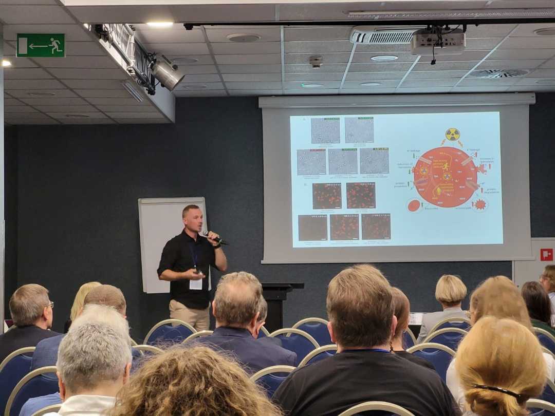 Sala wykładowa, ujęcie z tyłu sali uczestników siedzących na sali konferencyjnej podczas wykładu. Przed siedzącymi stojący prelegent przy prezentacji wyświetlanej z rzutnika.