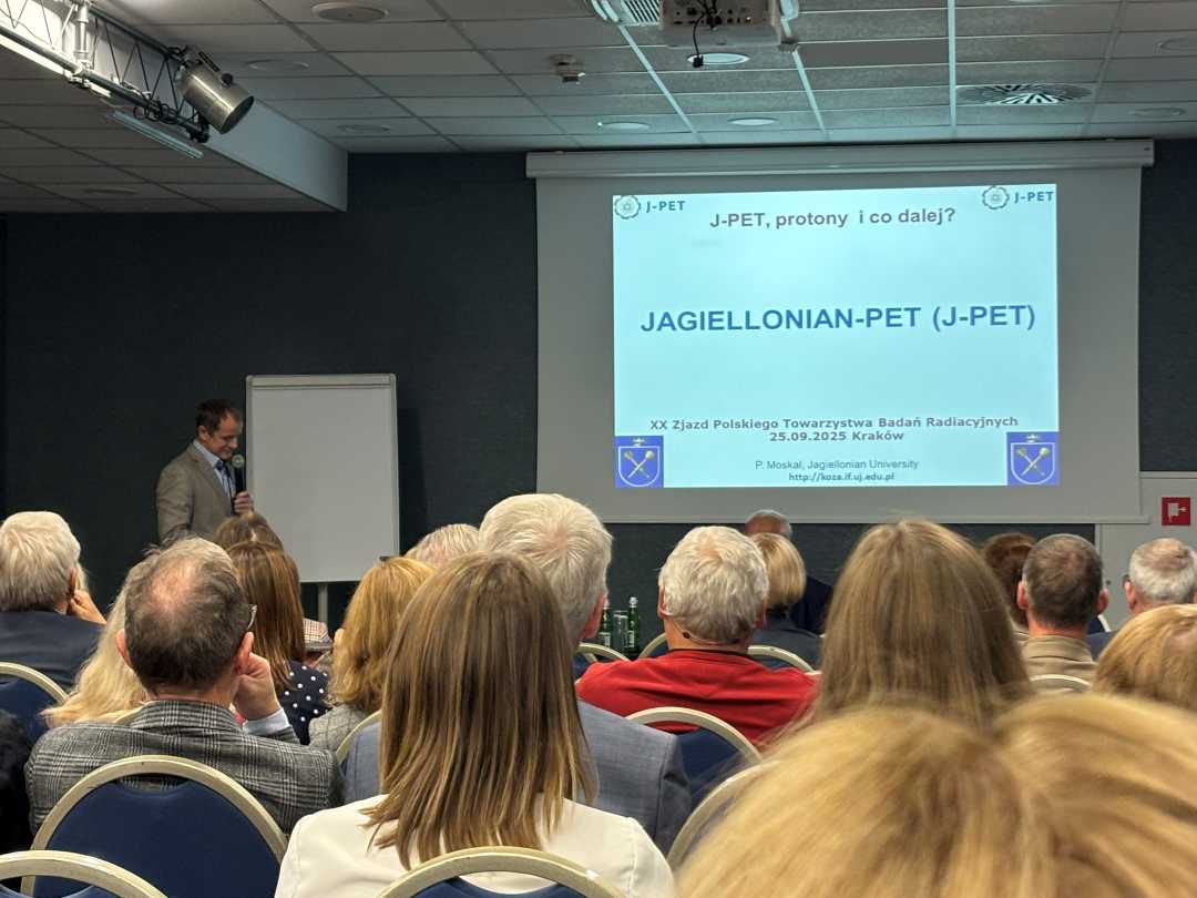 Sala wykładowa, ujęcie z tyłu sali uczestników siedzących na sali konferencyjnej podczas wykładu. Przed siedzącymi stojący prelegent przy prezentacji wyświetlanej z rzutnika.