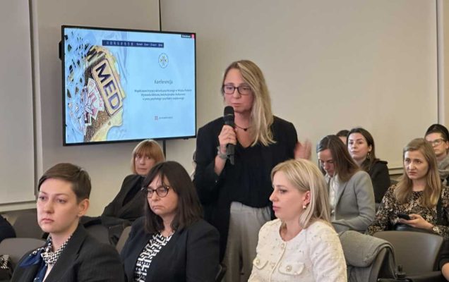 Kobieta na stoi sali konferencyjnej z stoi z mikrofonem w ręku, obok siedzący na krzesełkach słuchacze . Po jej prawej stronie wiszący ekran lcd z wyświetlaną prezentacji.