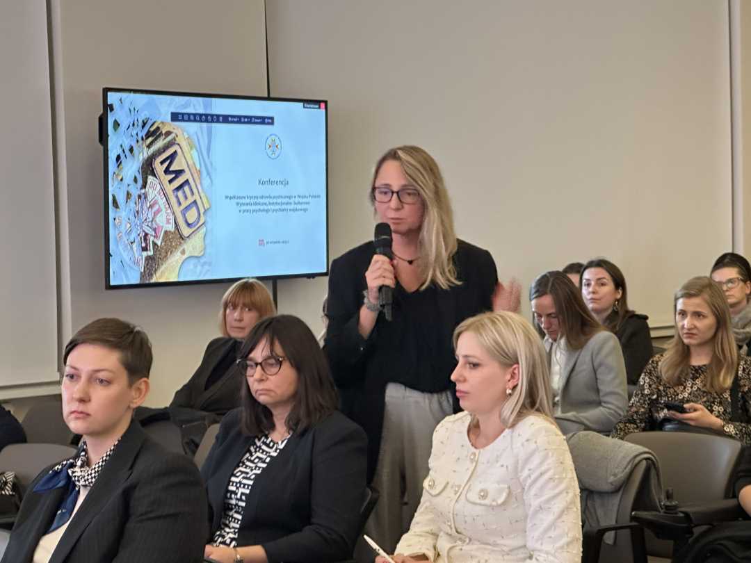 Kobieta na stoi sali konferencyjnej z stoi z mikrofonem w ręku, obok siedzący na krzesełkach słuchacze . Po jej prawej stronie wiszący ekran lcd z wyświetlaną prezentacji.