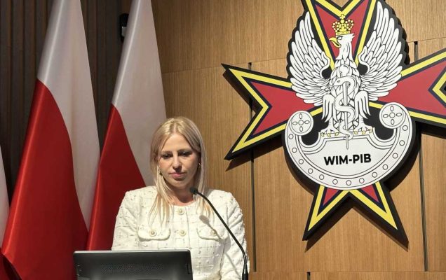 Prelegentka przy mównicy. Za jej plecami stoją flagi RP, na ścianie wisi logo WIM-PIB.