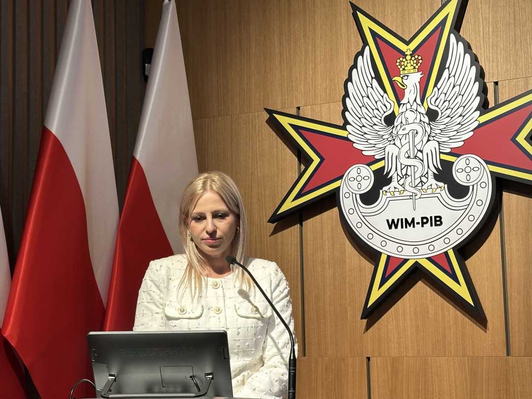 Prelegentka przy mównicy. Za jej plecami stoją flagi RP, na ścianie wisi logo WIM-PIB.