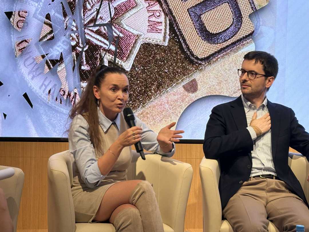 Prelegent i prelegentka siedzący w fotelach podczas panelu dyskusyjnego. Kobieta mówi do mikrofonu. Za ich plecami na dużym wyświetlaczu fragment prezentacji.