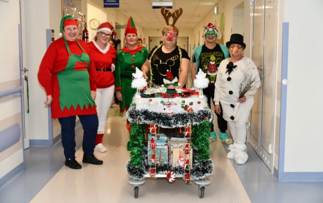 Pomocnicy świętego Mikołaja idą korytarzem szpitalnym z ozdobionym wózkiem pełnym prezentów.