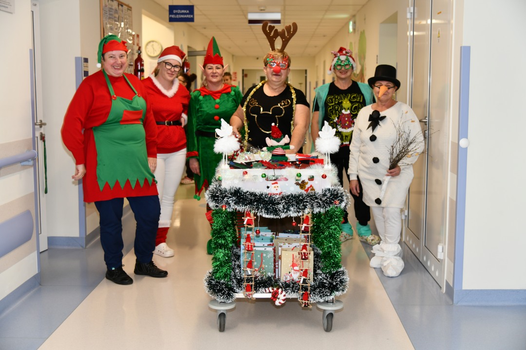 Pomocnicy świętego Mikołaja idą korytarzem szpitalnym z ozdobionym wózkiem pełnym prezentów.