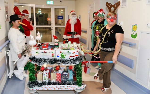 Pomocnicy świętego Mikołaja na korytarzu szpitalnym z ozdobionym wózkiem pełnym prezentów.