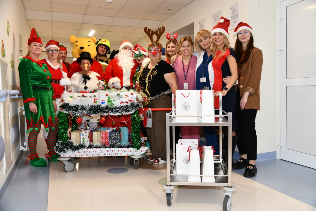 Pomocnicy świętego Mikołaja na korytarzu szpitalnym z ozdobionym wózkiem pełnym prezentów.