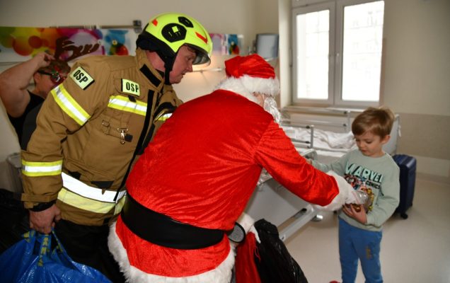 Święty Mikołaj i strażak na sali szpitalnej wręczają prezent kilkuletniemu chłopcu.