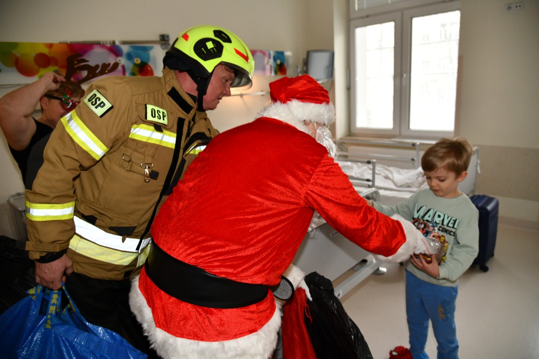 Święty Mikołaj i strażak na sali szpitalnej wręczają prezent kilkuletniemu chłopcu.