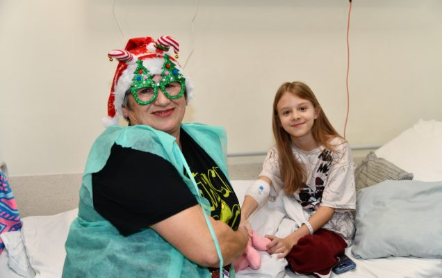 Pomocniczka świętego Mikołaja z kilkuletnią pacjentką siedzą na łóżku szpitalnym.