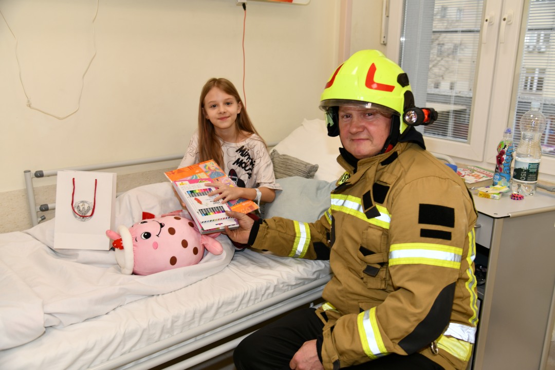 Sala szpitalna. Pan strażak wręcza prezent małej pacjentce.