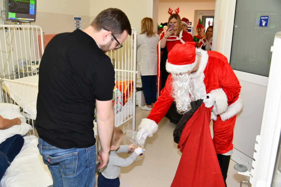 Sala szpitalna, święty Mikołaj pochyla się i wręcza 2 letniemu pacjentowi prezent. Za chłopcem stoi jego tata.