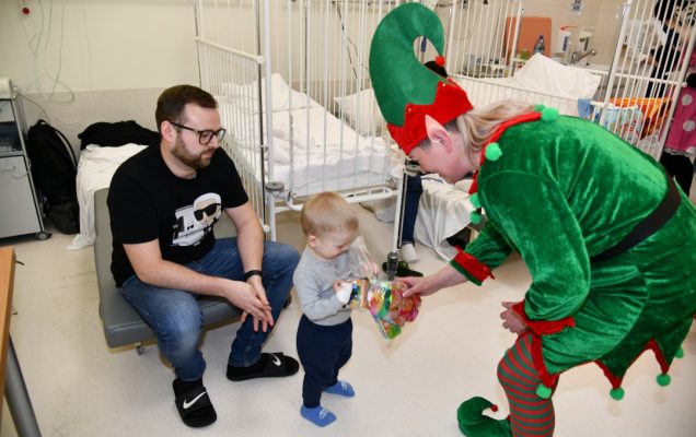 Sala szpitalna, Pani Elfowa pochyla się i wręcza 2 letniemu pacjentowi prezent. Za chłopcem kuca jego tata.