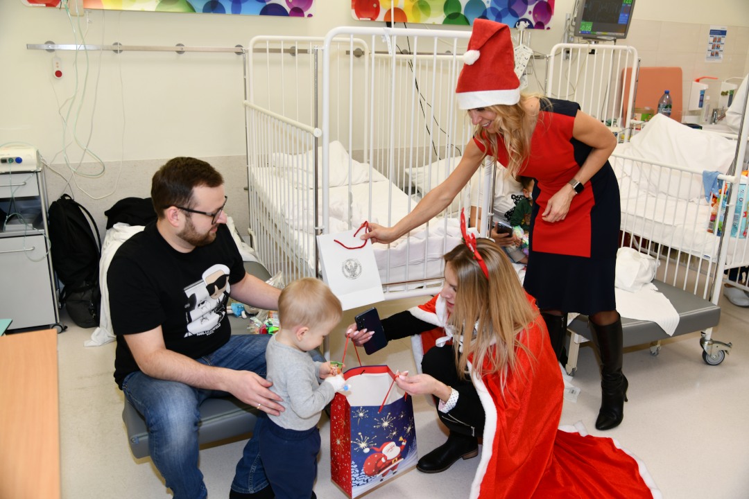 Sala szpitalna, Małgorzata Złotkowska pochyla się i wręcza 2 letniemu pacjentowi prezent. Za chłopcem stoi jego tata.