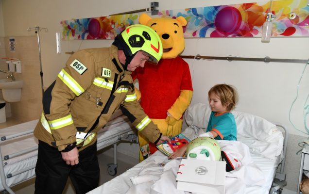 Sala szpitalna. Mały pacjent w łóżku szpitalnym ze strażakiem i Kubusiem Puchatkiem oglądają prezenty.