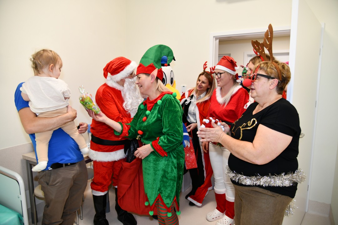 Święty Mikołaj i jego pomocnicy odwiedzają salę najmłodszych pacjentów. Tata z małym dzieckiem na ręku, jedna z pomocniczej wręcza dziecku prezent.