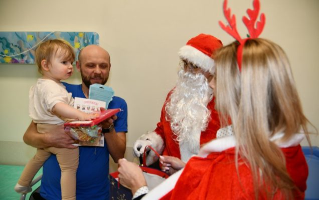 Święty Mikołaj i jego pomocnicy odwiedzają salę najmłodszych pacjentów. Tata z małym dzieckiem na ręku.
