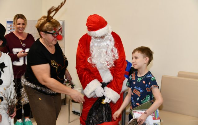 Święty Mikołaj w towarzystwie swoich pomocników daje prezent kilkuletniemu pacjentowi.