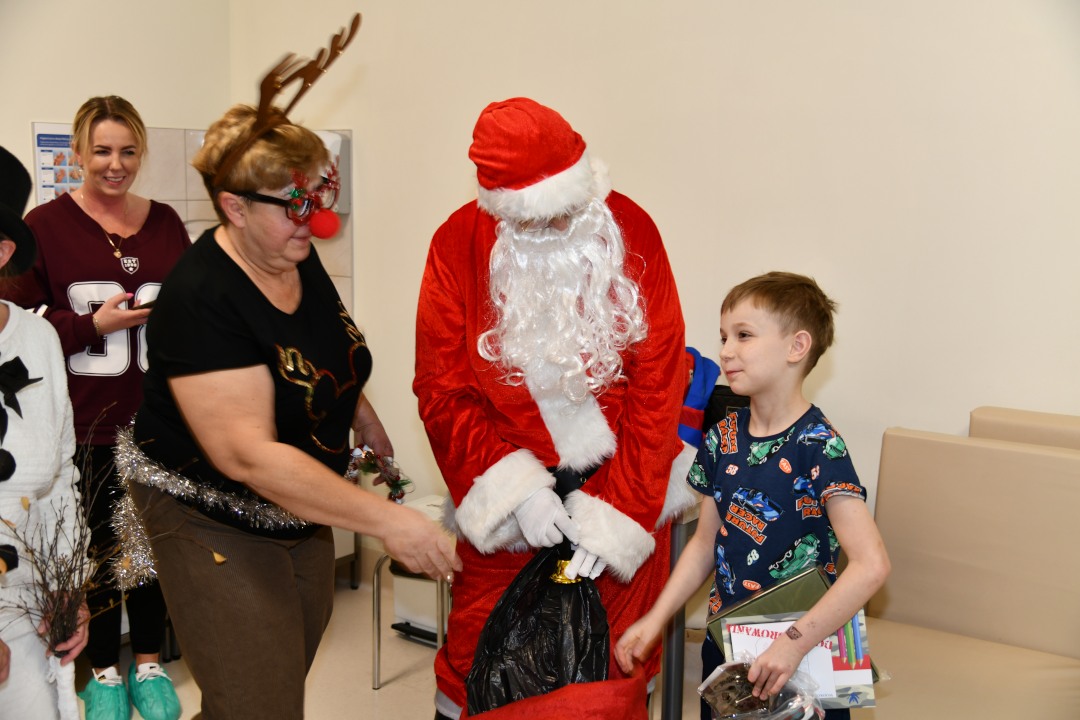 Święty Mikołaj w towarzystwie swoich pomocników daje prezent kilkuletniemu pacjentowi.