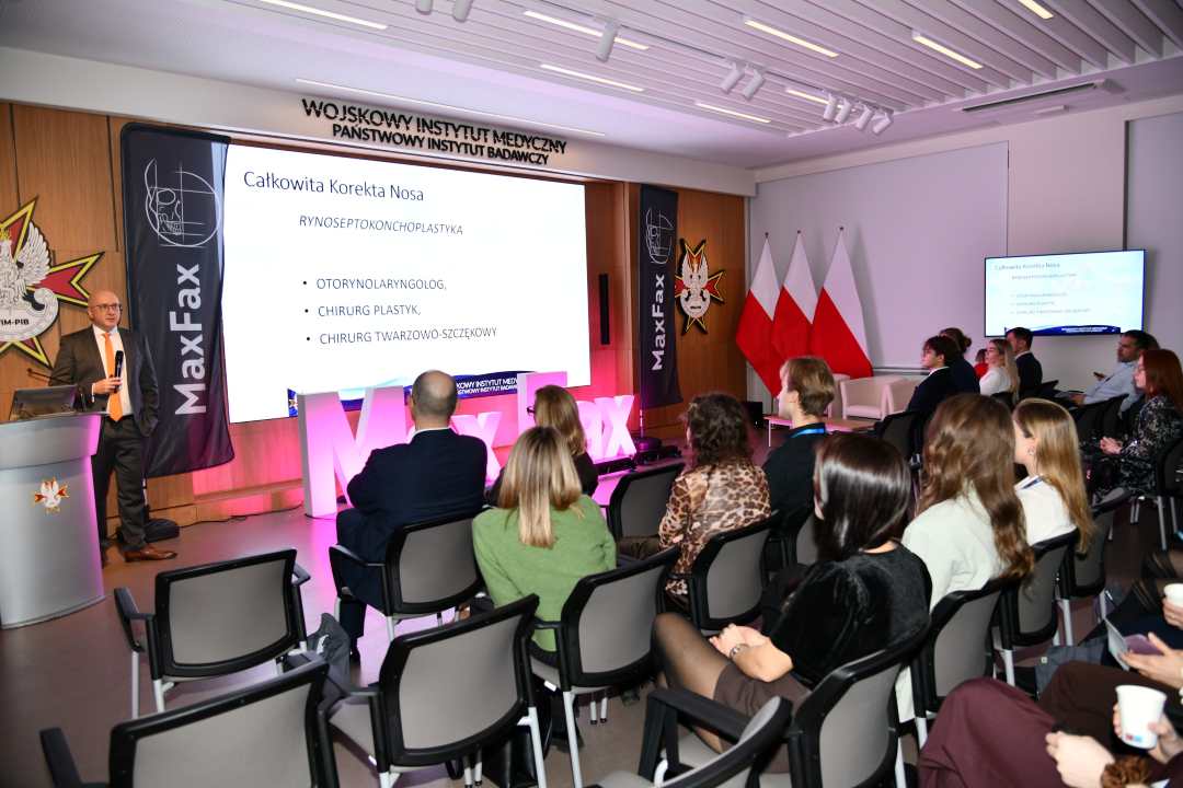 Siedzący słuchacze słuchający stojącego przed nimi prelegenta. Za prelegentem slajd z konferencji.