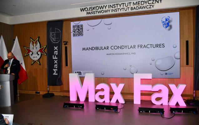 Prelegent przy mównicy. Obok niego stojący różowy napis Max Fax i ekran ze slajdem z konferencji.