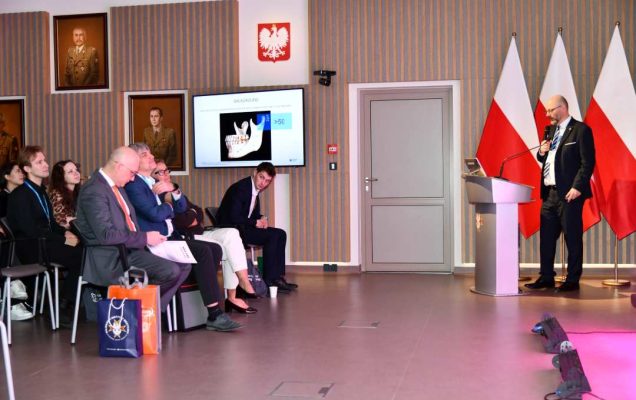 Prelegent przy mównicy mówiący do słuchaczy. Za nim logo WIM i stojące 3 flagi Polski.