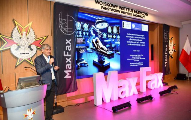 Prelegent przy mównicy. Za nim logo WIM. Obok stoi różowa dekoracja z napisem Max Fax i ekran z wyświetlonym slajdem z konferencji.