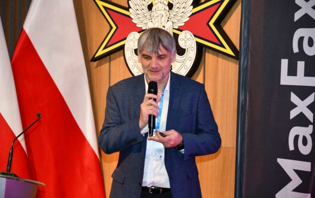 Prelegent z mikrofonem w ręku. Za nim logo WIM i flaga Polski, obok napis Max Fax.