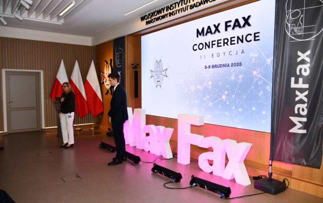 Prelegent podczas konferencji. Za nim podświetlony napis Max Fax i slajd tytułowy konferencji MAxFax.
