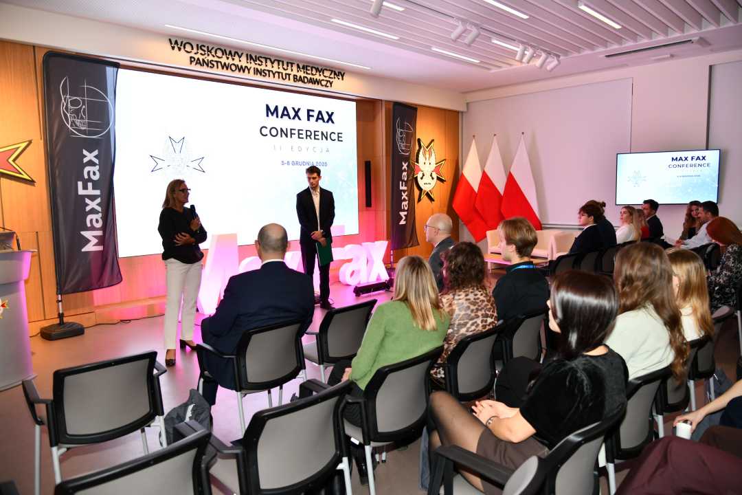 Doktor Aldona Chloupek z Prelegentem podczas konferencji. Za nimi podświetlony napis Max Fax i slajd tytułowy konferencji MAxFax. Przed nimi siedzący słuchacze konferencji.