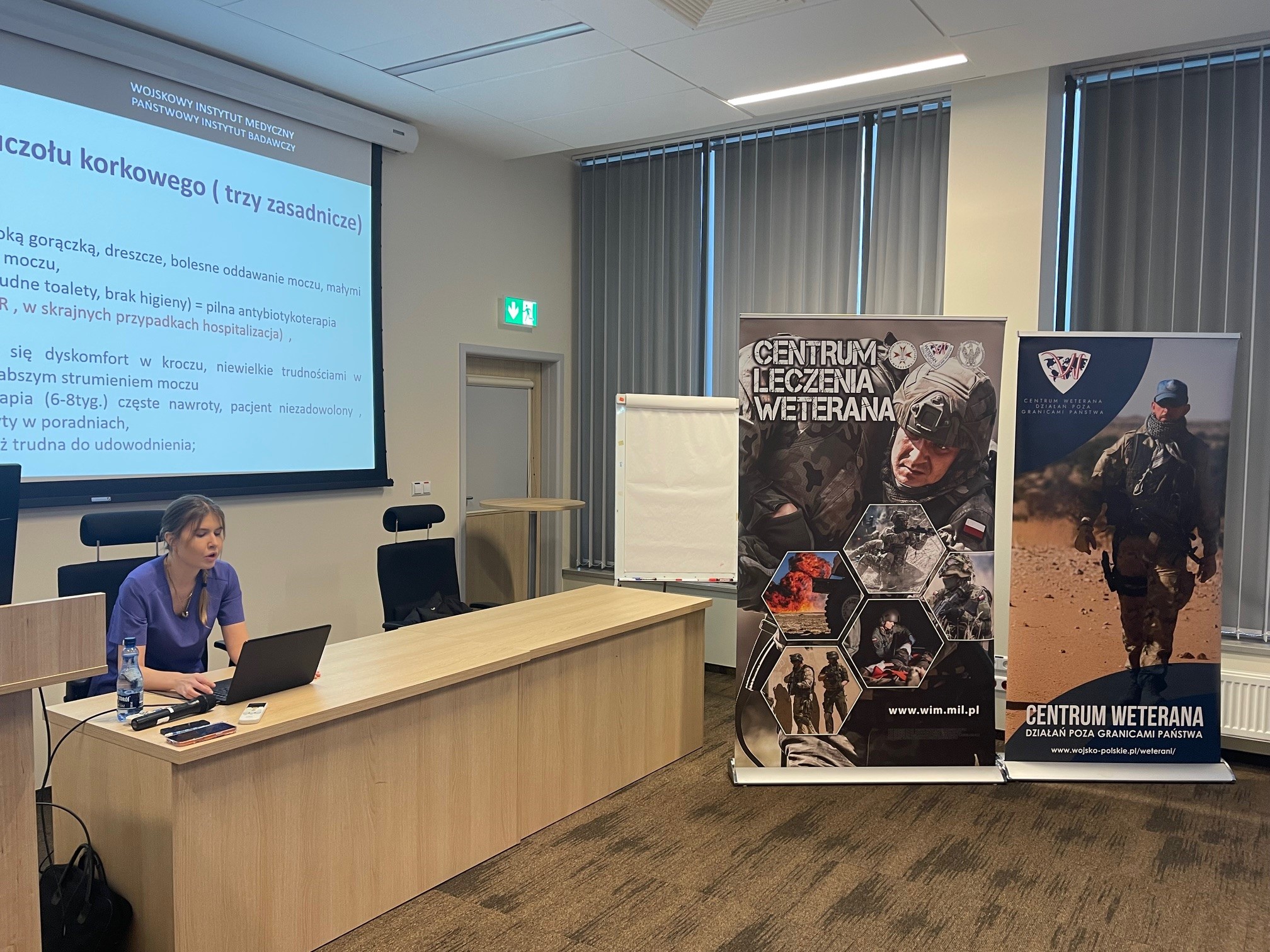 Ppłk dr n. med. Agnieszka Grabińska prowadzi wykład. Za jej plecami plansza wyświetlanej prezentacji. Z boku Stojak z plakatem Centrum leczenie weterana, na plakacie kolaż zdjęć z akcji wojskowych.