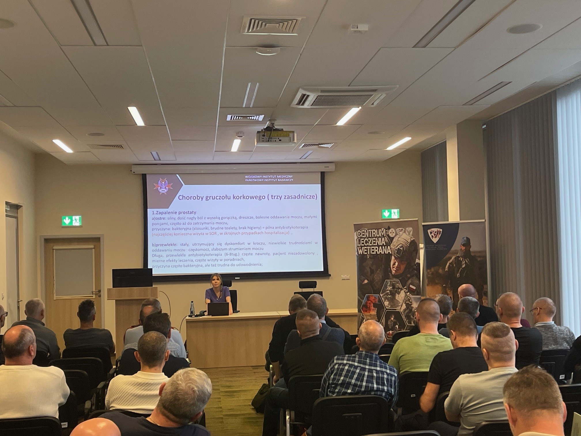 Ujęcie z tyłu na siedzących słuchaczy podczas wykładu. Ppłk dr n. med. Agnieszka Grabińska prowadzi wykład. Za jej plecami plansza wyświetlanej prezentacji. Z boku Stojak z plakatem Centrum leczenie weterana, na plakacie kolaż zdjęć z akcji wojskowych.