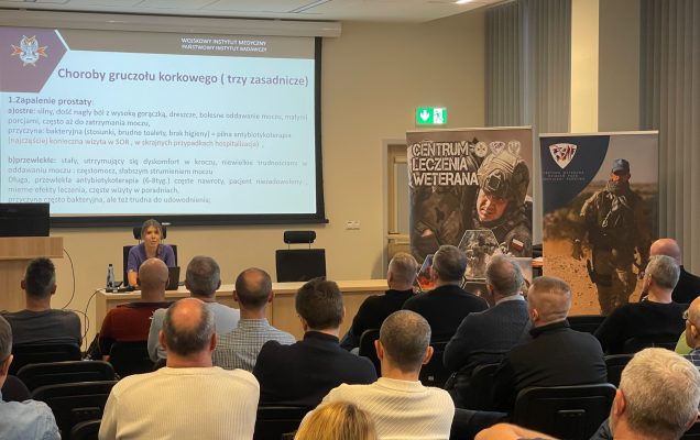 Ujęcie z tyłu na siedzących słuchaczy podczas wykładu. Ppłk dr n. med. Agnieszka Grabińska prowadzi wykład. Za jej plecami plansza wyświetlanej prezentacji. Z boku Stojak z plakatem Centrum leczenie weterana, na plakacie kolaż zdjęć z akcji wojskowych.