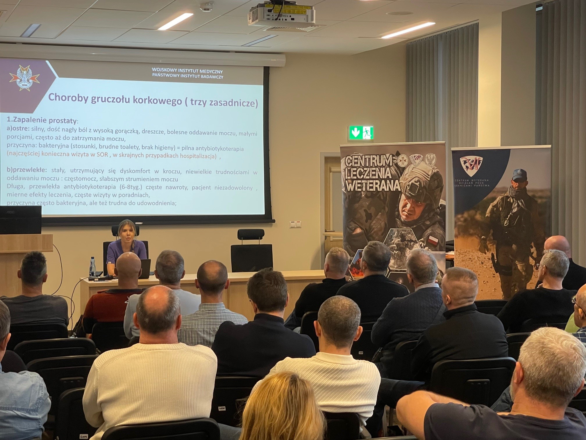 Ujęcie z tyłu na siedzących słuchaczy podczas wykładu. Ppłk dr n. med. Agnieszka Grabińska prowadzi wykład. Za jej plecami plansza wyświetlanej prezentacji. Z boku Stojak z plakatem Centrum leczenie weterana, na plakacie kolaż zdjęć z akcji wojskowych.