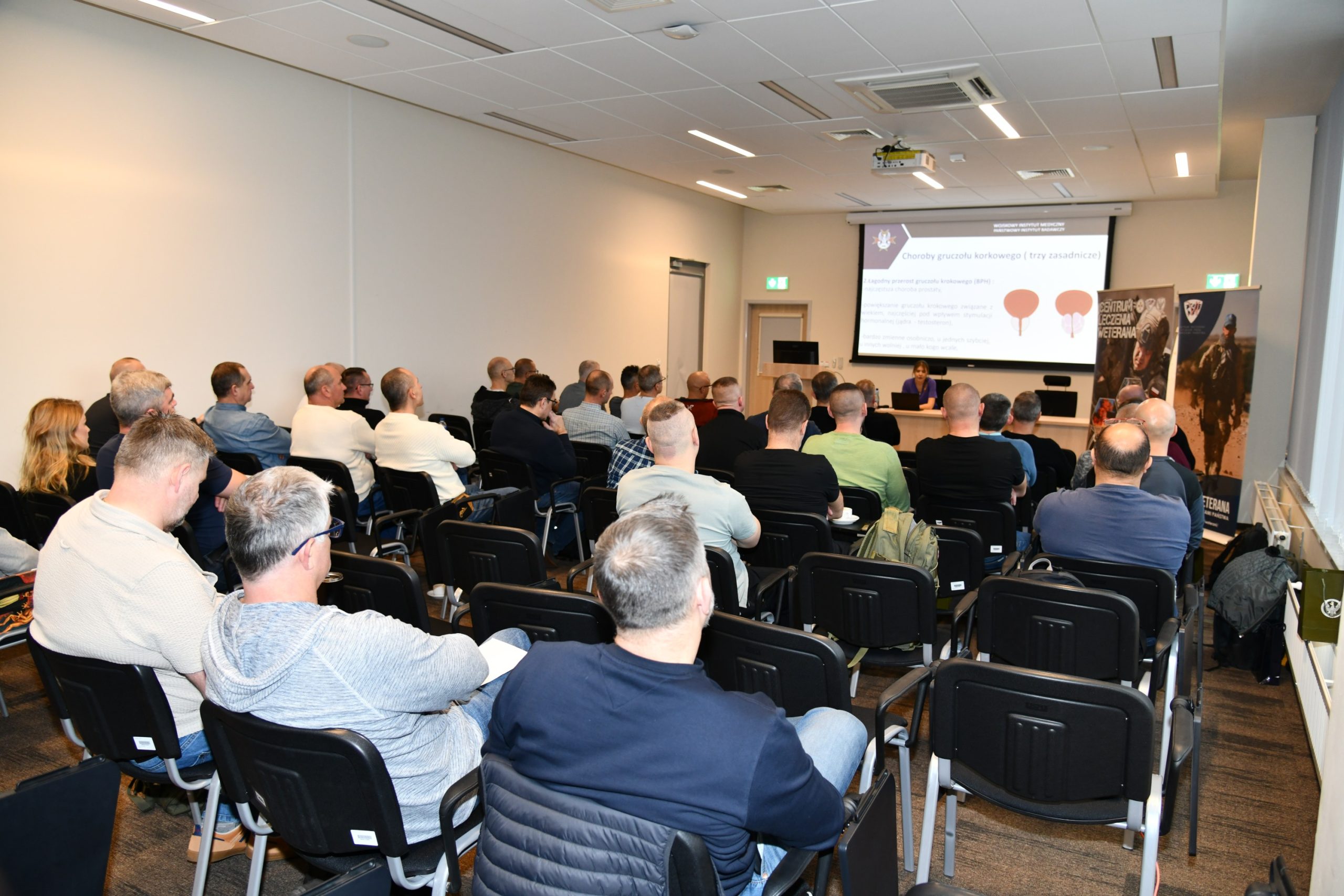 Ujęcie z tyłu na siedzących słuchaczy podczas wykładu. Ppłk dr n. med. Agnieszka Grabińska prowadzi wykład. Za jej plecami plansza wyświetlanej prezentacji. Z boku Stojak z plakatem Centrum leczenie weterana, na plakacie kolaż zdjęć z akcji wojskowych.