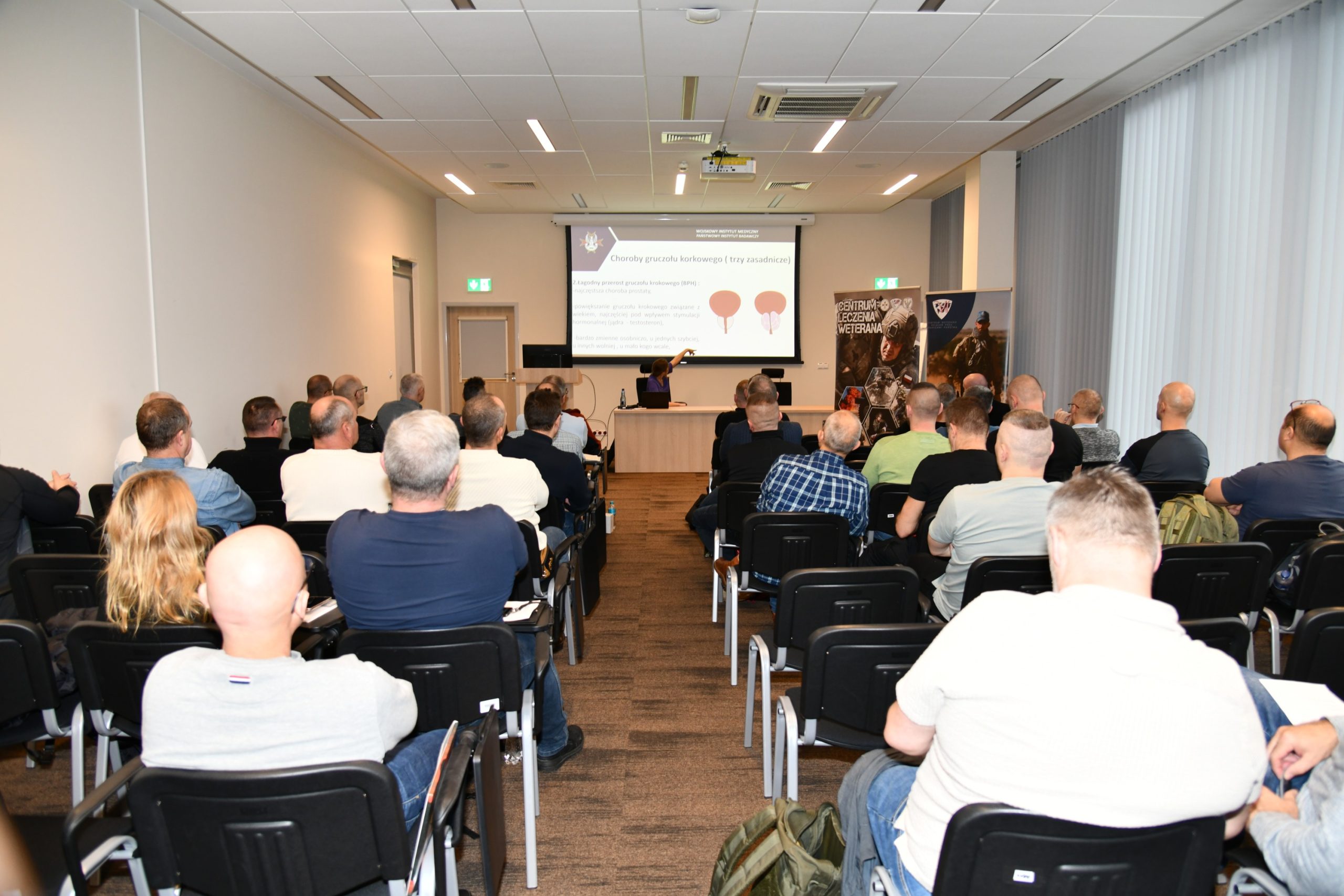 Ujęcie z tyłu na siedzących słuchaczy podczas wykładu. Ppłk dr n. med. Agnieszka Grabińska prowadzi wykład. Za jej plecami plansza wyświetlanej prezentacji. Z boku Stojak z plakatem Centrum leczenie weterana, na plakacie kolaż zdjęć z akcji wojskowych.