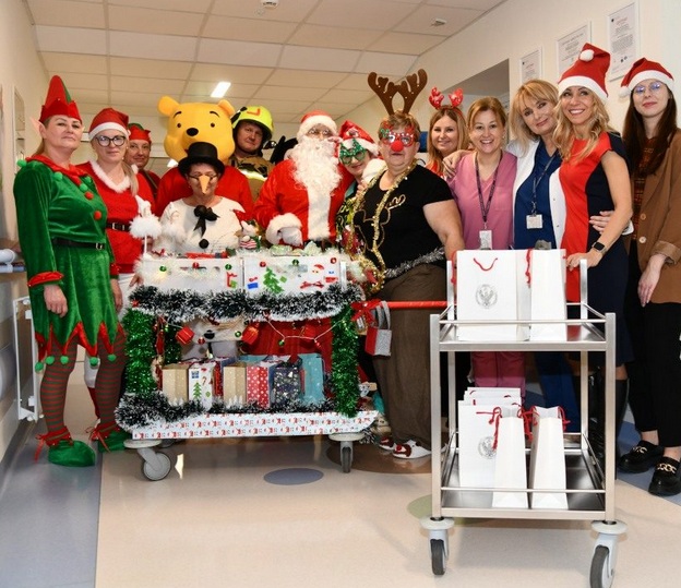 Pomocnicy świętego Mikołaja na korytarzu szpitalnym z ozdobionym wózkiem pełnym prezentów.