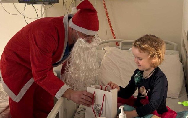 Święty Mikołaj pochyla się nad siedzącą małą dziewczynką na łóżku szpitalnej i wręcza jej torbę z prezentem.