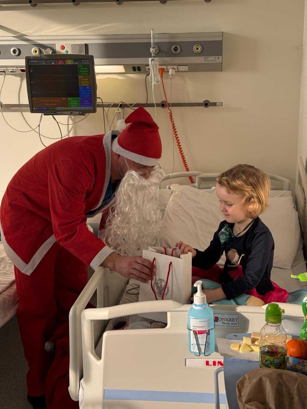 Święty Mikołaj pochyla się nad siedzącą małą dziewczynką na łóżku szpitalnej i wręcza jej torbę z prezentem.