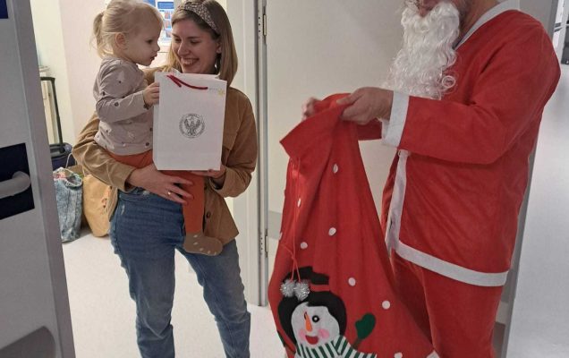 W drzwiach sali stoi uśmiechnięta mama z mała dziewczynką na rękach. Na przeciwko nich stoi Święty Mikołaj i wyjmuje prezent z worka.