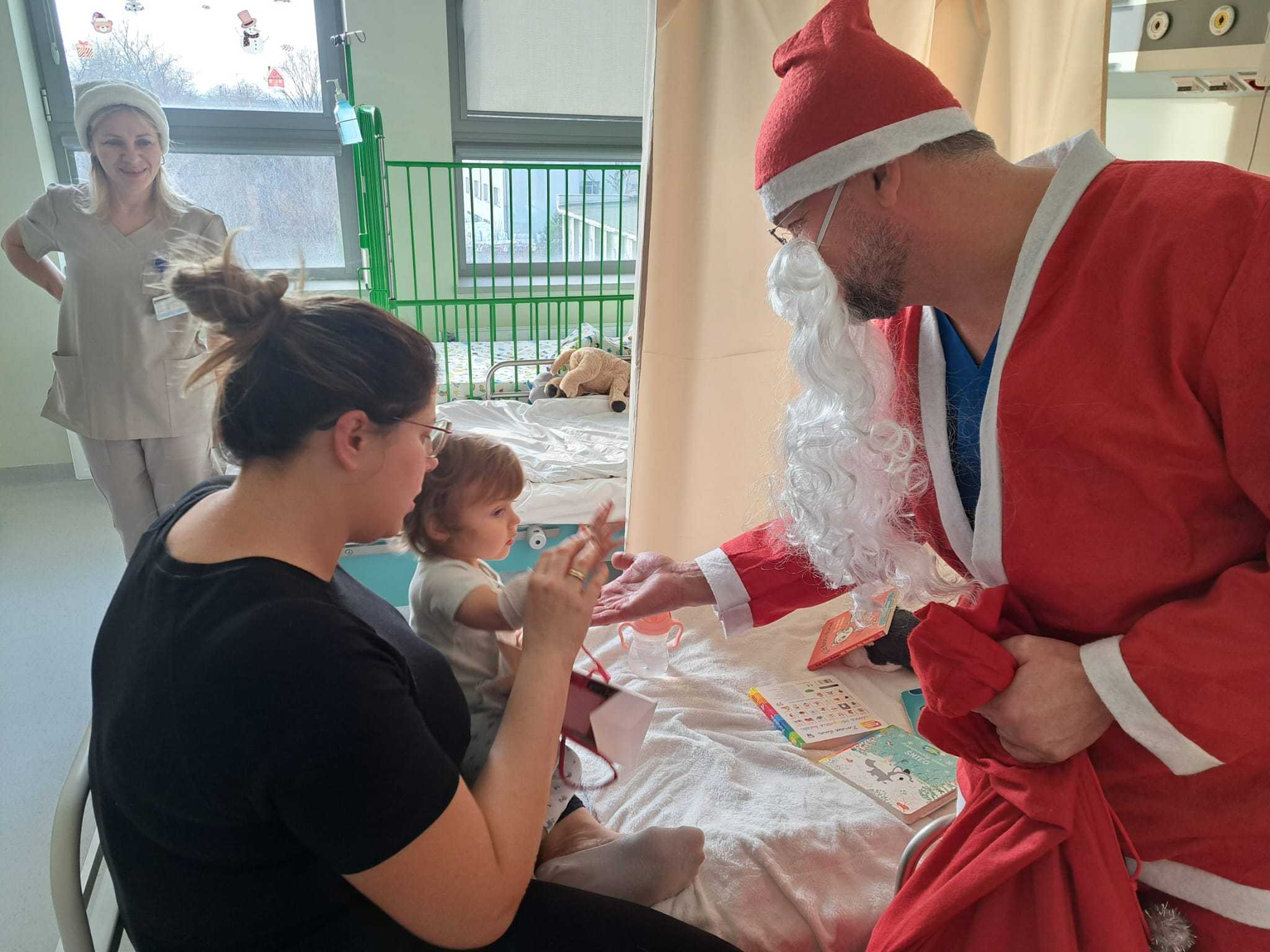 Święty Mikołaj pochyla się nad siedzącą małą dziewczynką i jej mamą przybija piątkę z dziewczynką.