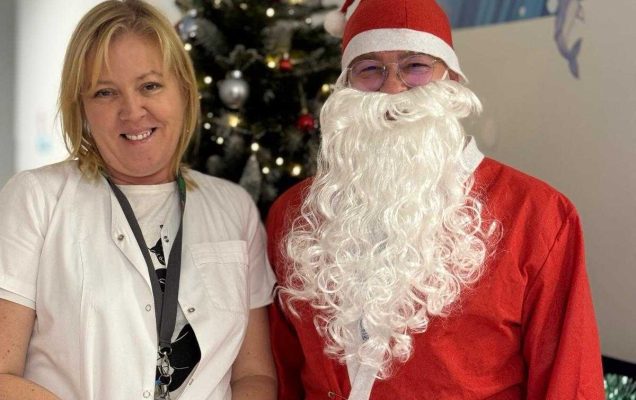 Święty Mikołaj pozuje na korytarzu szpitalnym z pracowniczką szpitala, z animi stoi udekorowana choinka.