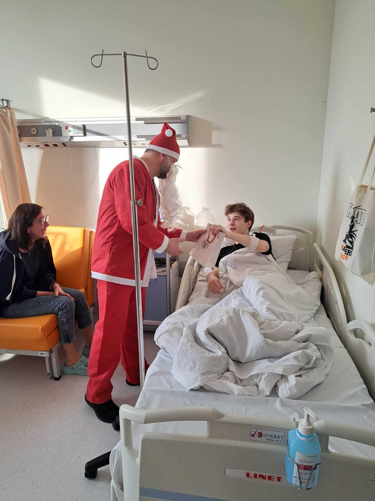 Święty Mikołaj pochyla się nad leżącym w łóżku szpitalnym nastolatkiem i wręcza mu torbę z prezentem.