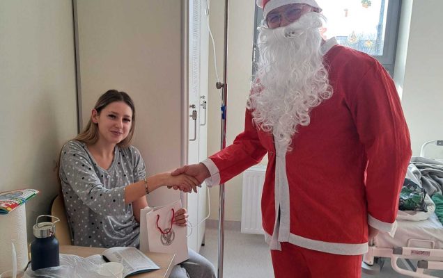 Święty Mikołaj pozuje do zdjęcia z siedzącą nastolatką w sali szpitalnej.