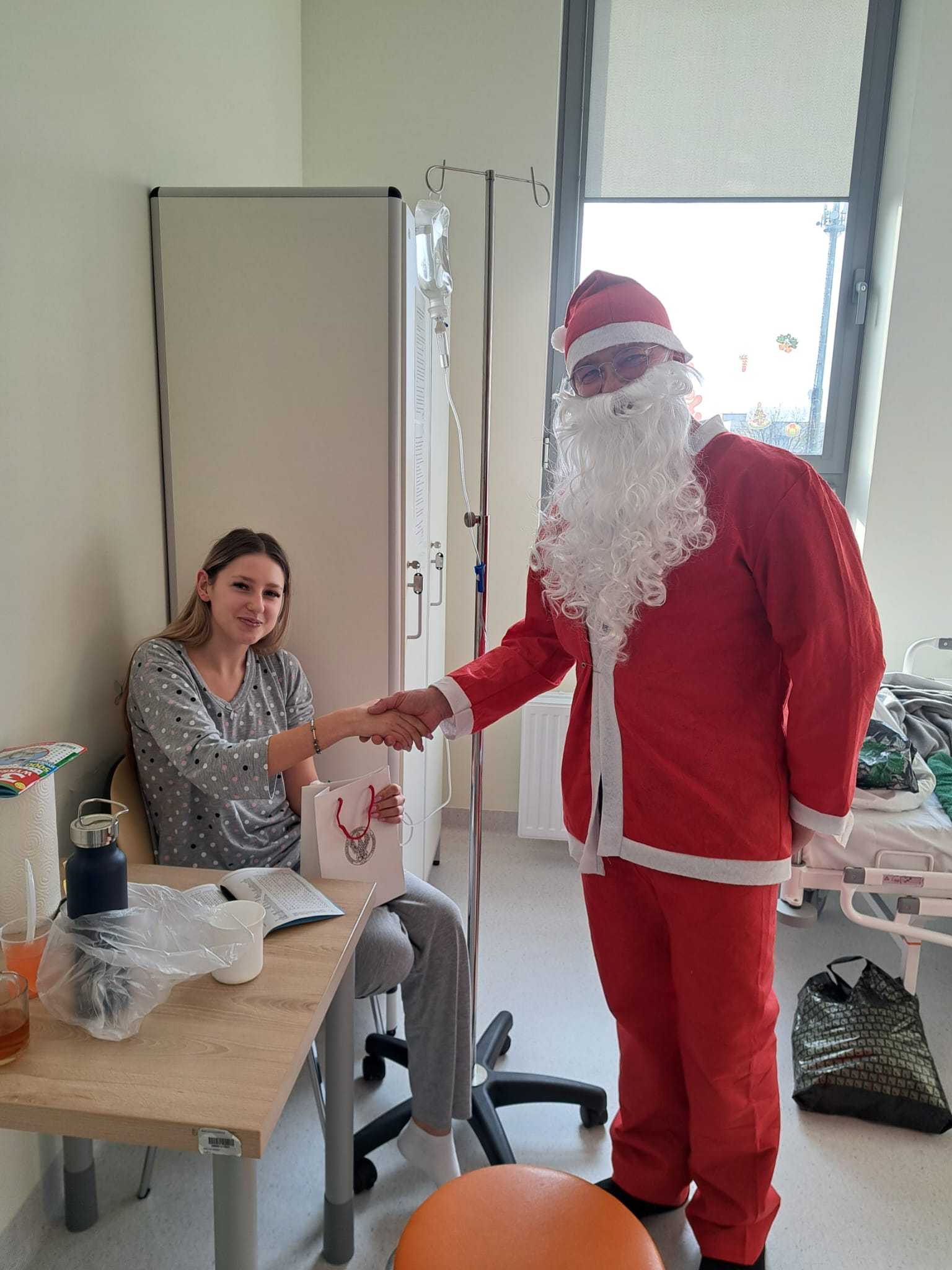 Święty Mikołaj pozuje do zdjęcia z siedzącą nastolatką w sali szpitalnej.